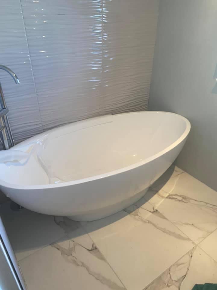 Freestanding oval bath installed on marble-effect floor tiles beside a textured white feature wall, with a floor-standing tap