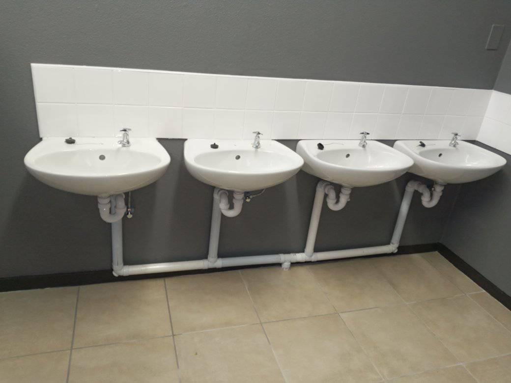 Row of four wall-hung basins with neatly run exposed pipework and traps along a tiled splashback